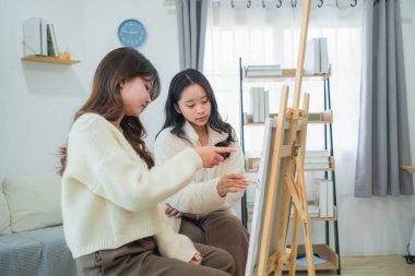 İki kadın kapalı bir stüdyoda sanat tekniklerini tartışıyor, resim sehpası ve doğal ışıkla yaratıcı ilhamı ve fikirleri sakince paylaşıyor.