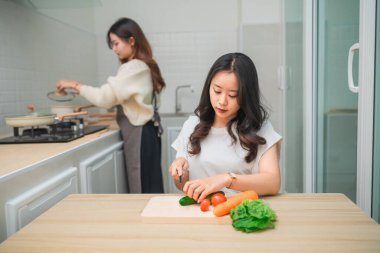 Kadınlar modern mutfakta sağlıklı yemekler hazırlarken, biri sebze doğrarken, diğeri taze malzemeler ve mutfak becerilerini ön plana çıkarıyor.