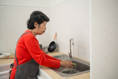 Kırmızı kazaklı yaşlı bir kadın mutfak lavabosunda sebze yıkıyor, evde sağlıklı yemek ve beslenmeyi teşvik ediyor, günlük hayatı ve yemek yapma alışkanlıklarını sergiliyor.