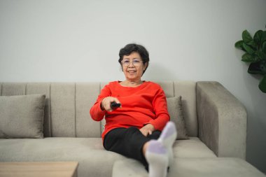 Neşeli yaşlı kadın kanepeye uzanıyor, uzaktan kumandayı tutuyor, evde boş vakitlerin tadını çıkarıyor, rahat bir yaşam alanında rahat kıyafetler giyiyor.