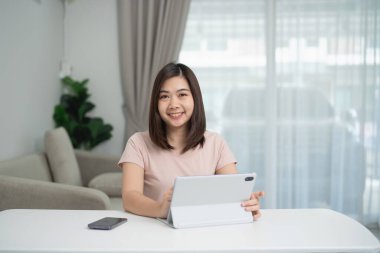 Genç bir kadın tablet cihazıyla masada oturuyor. Parlak modern oturma odasında neşeli bir gülümseme, doğal ışık ve yeşillikle iç içe.