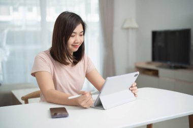 Evde dijital tablet kullanan genç bir kadın, modern oturma odasında doğal ışık ve yumuşak renklerle stilus ile öğrenmeye odaklanmış.