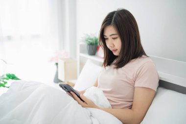 Yatakta otururken akıllı telefon kullanan genç bir kadın. Yumuşak sabah ışığıyla rahat bir atmosfer. Kapalı alandaki tesisin rahatlamasını sağlıyor.