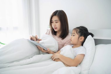 Anne ve kızı, rahat bir yatak odasında, yumuşak doğal ışık altında kitap okurken birlikte güzel vakit geçiriyorlar.