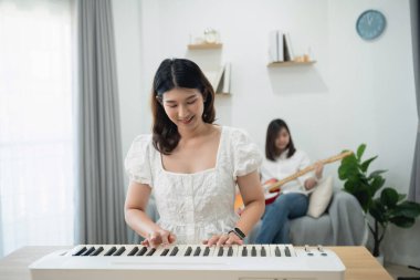 Neşeli genç bir kadın parlak oturma odasında gitar çalan bir arkadaşıyla klavye çalıyor, müzik keyfi ve ev ortamında yaratıcı arkadaşlıklar sergiliyor.