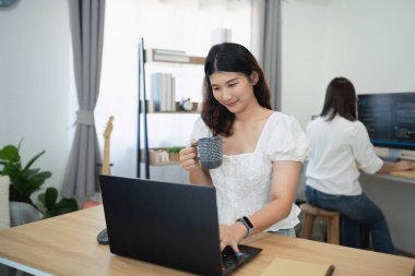 Bilgisayarda, modern ev ofisinde kahve fincanı ile çalışan genç bir kadın, arka planda iş arkadaşı, gündelik ve şık çalışma alanı ayarlarıyla ekrana odaklanıyor.