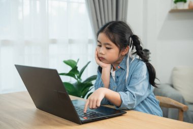 Evde online öğrenim için dizüstü bilgisayar kullanan sıkıcı Asyalı kız. Yorgun çocuk masada oturuyor, stresli ya da ev ödevlerinde tembellik ediyor, bilgisayar başında çalışıyor..