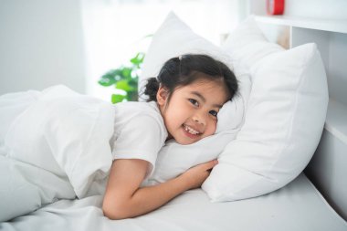 Neşeli genç kız, yumuşak bir yastıkta uzanırken gülümsüyor. Parlak bir yatak odasında, doğal ışıkla rahat ve davetkar bir atmosfer yaratıyor.