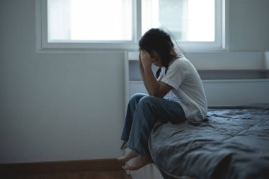 Yatakta oturan genç bir kadın, üzgün ve sıkıntılı hissediyor, elleriyle yüzünü kapatıyor, iç mekan ayarları, pencereden gelen doğal ışık, duygusal bir an.
