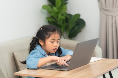 Genç kız evde çalışırken dizüstü bilgisayara odaklandı, günlük giysiler giydi ve rahat oda ortamında özenle dijital cihazlar üzerinde çalıştı.