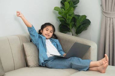 Dizüstü bilgisayarı olan neşeli bir çocuk kutlama için yumruğunu kaldırıyor, rahat bir kostüm giyiyor, arka planda yeşil bitkiler olan kapalı bir ortamda.