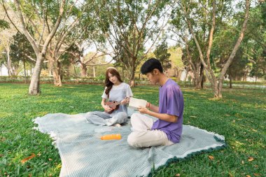 Genç çift sakin bir parkta, ağaçların arasından süzülen yemyeşil ve güneş ışığıyla çevrili, ukulele oynayıp kitap okumaktan keyif alıyor..