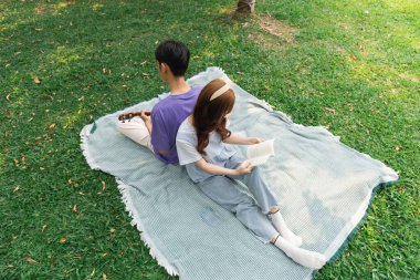 Genç bir çift parkta piknik battaniyesinde oturup dinlenmekten keyif alıyor. Etrafı otlarla çevrili, huzurlu bir ortamda kitap okuyor ve dinleniyor.