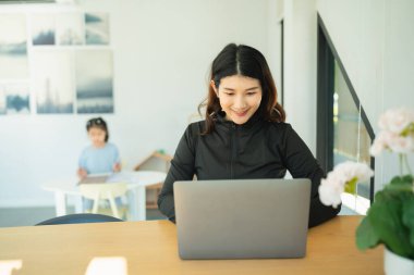 Modern ev ofisinde dizüstü bilgisayarda çalışan gülümseyen bir kadın, arka planda, parlak ve rahat iç mekanlarda okuyan bir çocuk, üretkenlik ve aile zamanına odaklanıyor