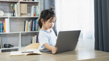 Evde online öğrenim gören küçük bir çocuk, dizüstü bilgisayara odaklanmış, rahat ve parlak bir odada ahşap bir masada oturuyor, çocuklar için eğitici aktiviteler yapıyor.