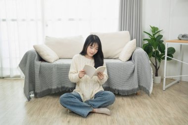 Rahat bir kanepe ve çiçekli, rahat bir oturma odasında oturmuş kitap okuyan bir kadın, bir anlık rahatlama ve iç huzurun tadını çıkarıyor.