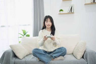 Genç bir kadın rahat koltuğunda oturmuş modern şık oturma odasında kontrolörle video oyunu oynuyor. Rahat atmosferinin tadını çıkarıyor.