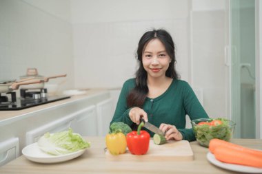 Parlak bir mutfakta taze sebzeler hazırlayan genç bir kadın, evde besleyici bir yemek için renkli malzemelerle sağlıklı aşçılık pratiği yapıyor.