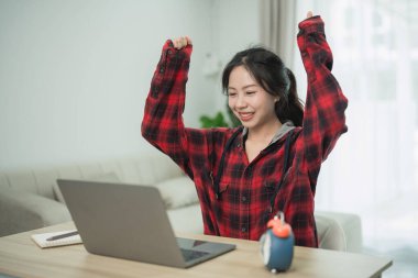 Mutlu genç kadın, iç mekanda rahat bir laptopta çalışırken, fanila gömleği giyerken ve ahşap masada otururken başarıyı kutluyor.