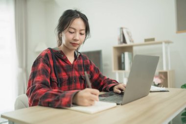 Genç bir kadın dizüstü bilgisayarla modern ev ofisinde çalışırken deftere bir şeyler yazıyor. Yaratıcı zamanlarının tadını çıkarıyor. Gündüz kıyafetleri, rahat ortamları.