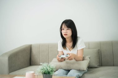 Kanepede oturan genç bir kadın elinde bir kontrolörle video oyunu oynuyor. Modern bir yaşam alanında boş zamanların tadını çıkarıyor.