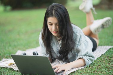 Çevresi yeşilliklerle çevrili, huzurlu bir park ortamında dizüstü bilgisayarda çalışan genç bir kadın, gündüz vakti verimlilik ve rahatlama için boş vakitlerin tadını çıkarıyor.