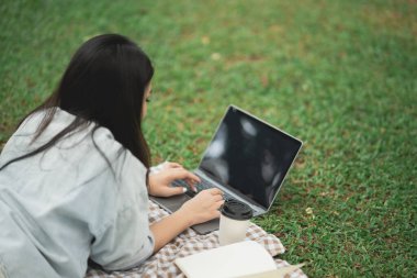 Genç bir kadın dışarıda çimenlikte kahve fincanı ile dizüstü bilgisayarda çalışıyor, boş zamanlarında huzurlu doğa ortamının tadını çıkarıyor, görevine odaklanıyor.