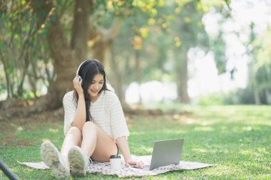 Genç bir kadın dışarıda dizüstü bilgisayarla müzik dinlerken Sunny Park 'ta çimlerde oturuyor kulaklık takıyor ve rahat kıyafetler giyiyor.