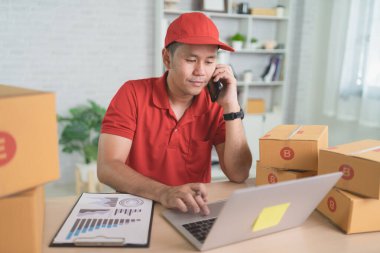 Kırmızı şapkalı odaklanmış bir adam, parlak bir ofis yerinde, karton kutular ve evrak işleriyle dolu bir masada laptopuyla çalışırken telefonla konuşuyor..