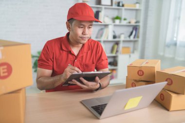 Kırmızı üniformalı bir teslimatçı, karton kutularla dolu bir masada sevkiyat detaylarını incelerken yoğun bir lojistik ortamı gösteriyor..
