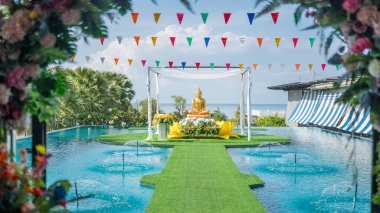 Serene Outdoor Ceremony with Golden Buddha Statue Surrounded by Lush Greenery and Festive Decorations by Poolside Under Clear Blue Sky