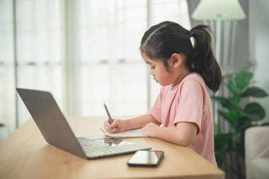 Çalışma ve yaratıcılık ile meşgul olan genç kız, dizüstü bilgisayarlı ve akıllı telefonlu notlar yazıyor, kapalı alanda doğal ışık, odaklanmış öğrenme atmosferi