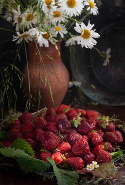 Bir demet taze çilek ve yabani papatyalarla dolu kır kil sürahisi. Ahşap masa üzerinde koyu arka plan, seçici odak, dikey, seçici odak, sade anahtar..