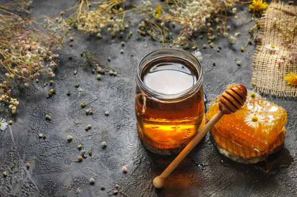 Bal peteği ve şeffaf bal koyu arka planda bir kavanozda kuru ve taze çiçeklerle. Doğal ışıklandırmada seçici yumuşak odak. Metin için alanı kopyala