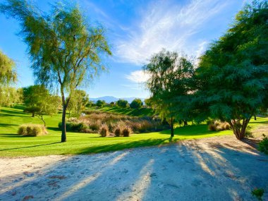 Çöldeki vaha sahnesinde yemyeşil ağaçlar ve parlak mavi gökyüzü ile uzaktaki dağlar gibi derin gölgeler ve parlak güneş ışınları bir golf sahası ya da tatil köyü gibi