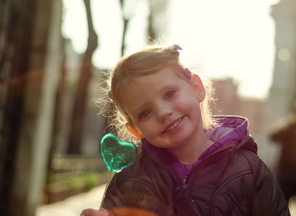 Cute little girl with heart shaped candy smiling in camera — Stock Photo, Image