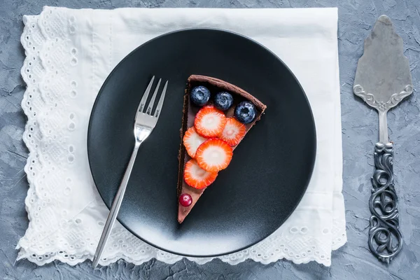 Piece of chocolate pie (cake, tart) decorated with fresh fruits and berries on black plate
