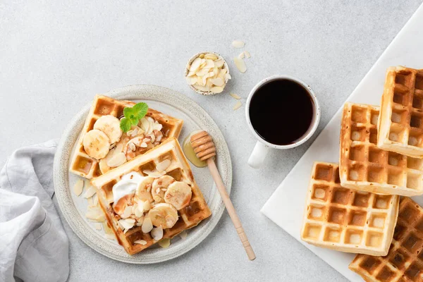 Sweet banana belgian waffles and coffee. Square waffles with banana, cream, almond nuts and caramel sauce and cup of black coffee on grey concrete background, top view
