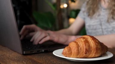 Kafede dizüstü bilgisayarda çalışan ve kruvasan yiyen serbest çalışan bir kadın. Mesafe işi, klavyede yazan hippi kadın.