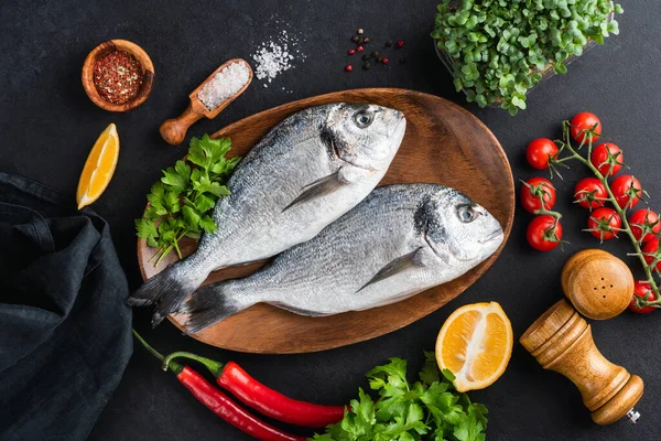 Dorado fish with vegetables and spices ready for cooking, table top view, black concrete background. Two raw uncooked dorado sea fish on wooden tray, healthy cooking process