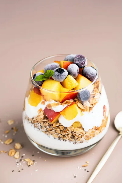 Greek yogurt, granola and fruits in jar. Healthy vegetarian breakfast parfait with peach, blueberries, oat honey granola and yogurt, beige background