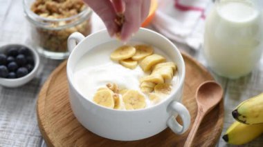 Yoğurt kasesine granola ekliyorum. Muz dilimli Yunan yoğurdu, gevrek yulaf ezmesi.