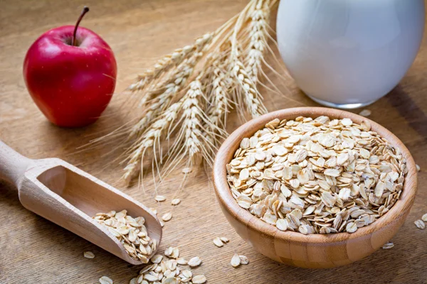 Oatmeal (oat flakes, rolled oats), fresh milk, wheat ears and apple on wooden texture background — Stock Photo, Image