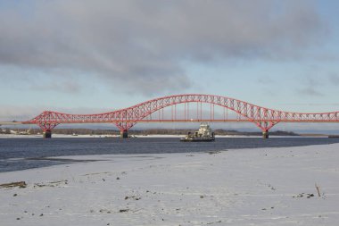 Ugra 'daki köprünün arka planına karşı bir römorkör. Khanty-Mansiysk 'teki Kızıl Ejder Köprüsü' nün arka planında karlı kıyıları olan bir nehirde römorkör.