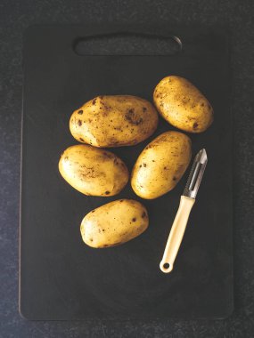 Patatesler ve arka planda gri mutfak tezgahı olan siyah bir tahta üzerinde soyulmuş bir bıçak..