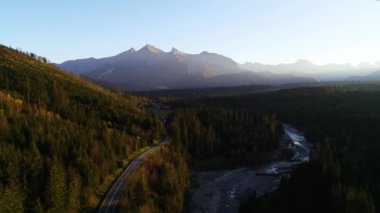Dağlık yolun havadan görünüşü. Dağlarda güzel, ılık bir sonbahar ve renkli ormanda güneşli, temiz bir yol. Vahşi dağ manzarasında otobanın destansı drone görüntüleri.