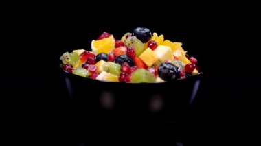 Kasedeki renkli meyve salatası 4K 'de siyah arka planda dönüyor. Taze ve lezzetli salatayı portakal, elma, böğürtlen, nar, kivi, ananas, ahududu, yabanmersini ve çilekle kapatın.