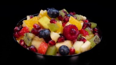 Kasedeki renkli meyve salatası 4K 'de siyah arka planda dönüyor. Taze ve lezzetli salatayı portakal, elma, böğürtlen, nar, kivi, ananas, ahududu, yabanmersini ve çilekle kapatın.