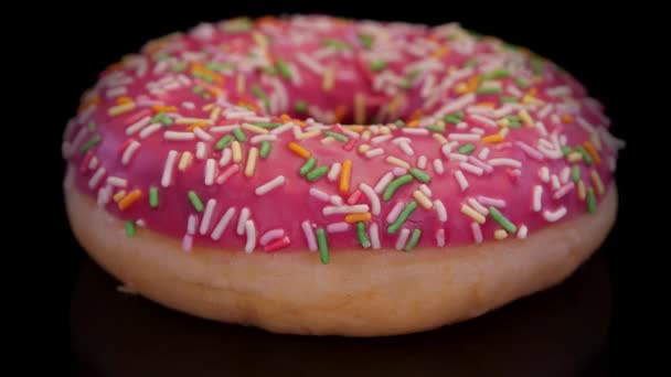 带粉红糖霜的甜甜圈 色彩艳丽的洒落在黑色的背景上 近距离观察 甜甜甜圈与霜冻和撒布隔离 垃圾食品 — 图库视频影像
