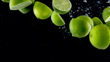 Limon yavaş çekimde suya sıçrıyor. Siyah arka planda yavaş yavaş düşen bir sürü limon. Siyah arka planda izole edilmiş organik yeşil turunçgil. Meyveli taze su.